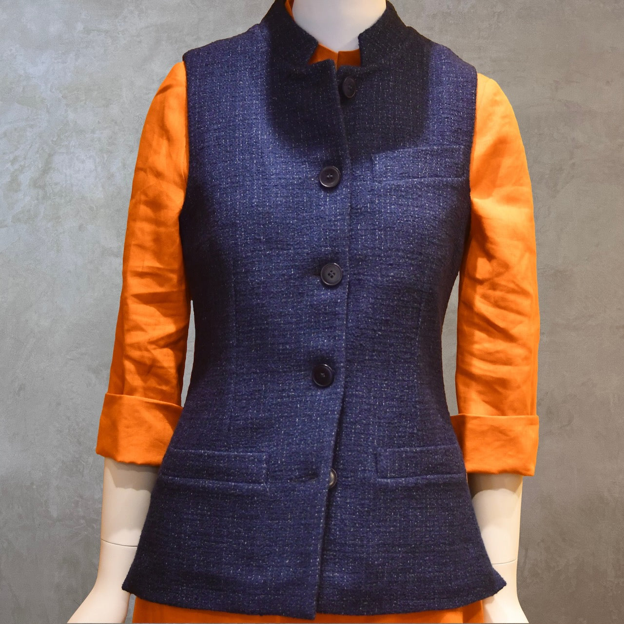 Ladies Shaped Gilet in Linton Tweeds boucle. A deep navy spring-summer weight fabric, this gilet packs superbly and can be worn everywhere. Handmade in London. Front Shot shown over orange lightweight linen dress from EdNerat.
