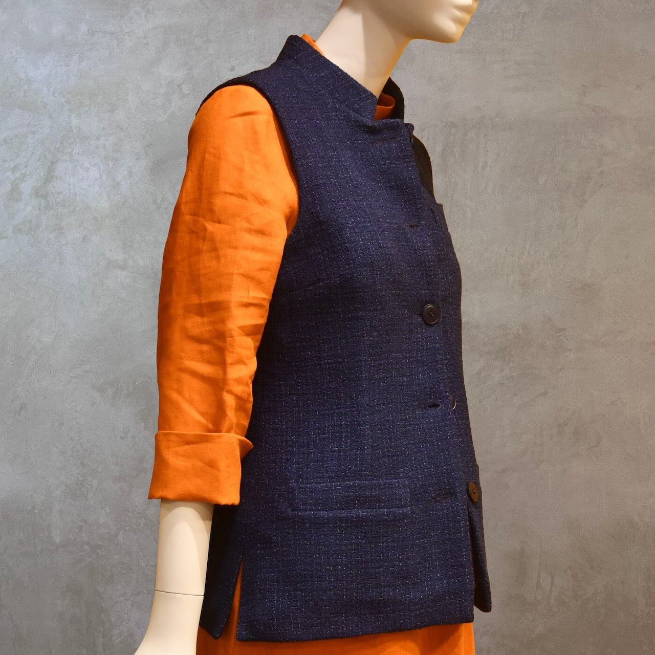 Ladies Shaped Gilet in Linton Tweeds boucle. A deep navy spring-summer weight fabric, this gilet packs superbly and can be worn everywhere. Handmade in London. Angled Side Shot shown over orange lightweight linen dress from EdNerat.