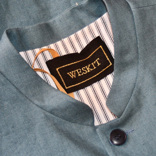 Close up of label on Light blue loose cut linen gilet from Weskit. Handmade in London, this slubby linen waistcoat is made in pure Irish linen and lined in striped cotton ticking. Mandarin collar and Courtney and Co buttons. Entirely made in Great Britain.