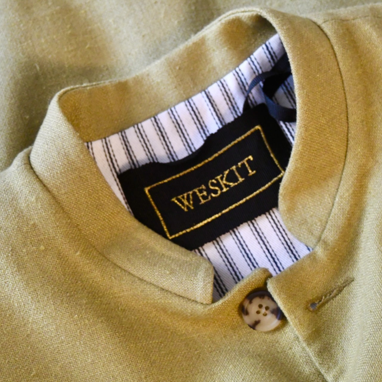 Relaxed fit summer weight gilet in green silk weave. Handmade in London this gilet has a Mandarin collar and is fully lined in pure cotton ticking. Made in Great Britain. Close up shot showing mandarin collar, black silk embroidered label and striped cotton ticking lining.