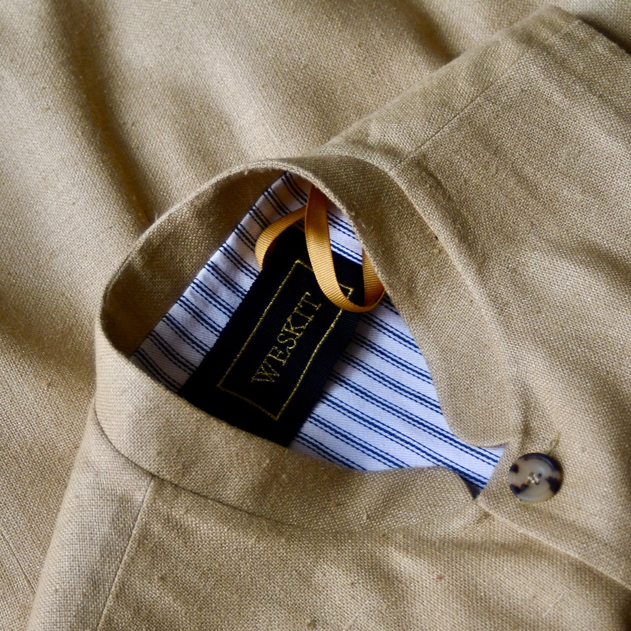 Close up on collar of lighter weight fitted gilet in sandy straw coloured silk weave fabric. Handmade in London this Nehru style gilet boast a Nehru collar, five pockets and is fully lined in pure cotton ticking. Detail shot shows Mandarin Collar.