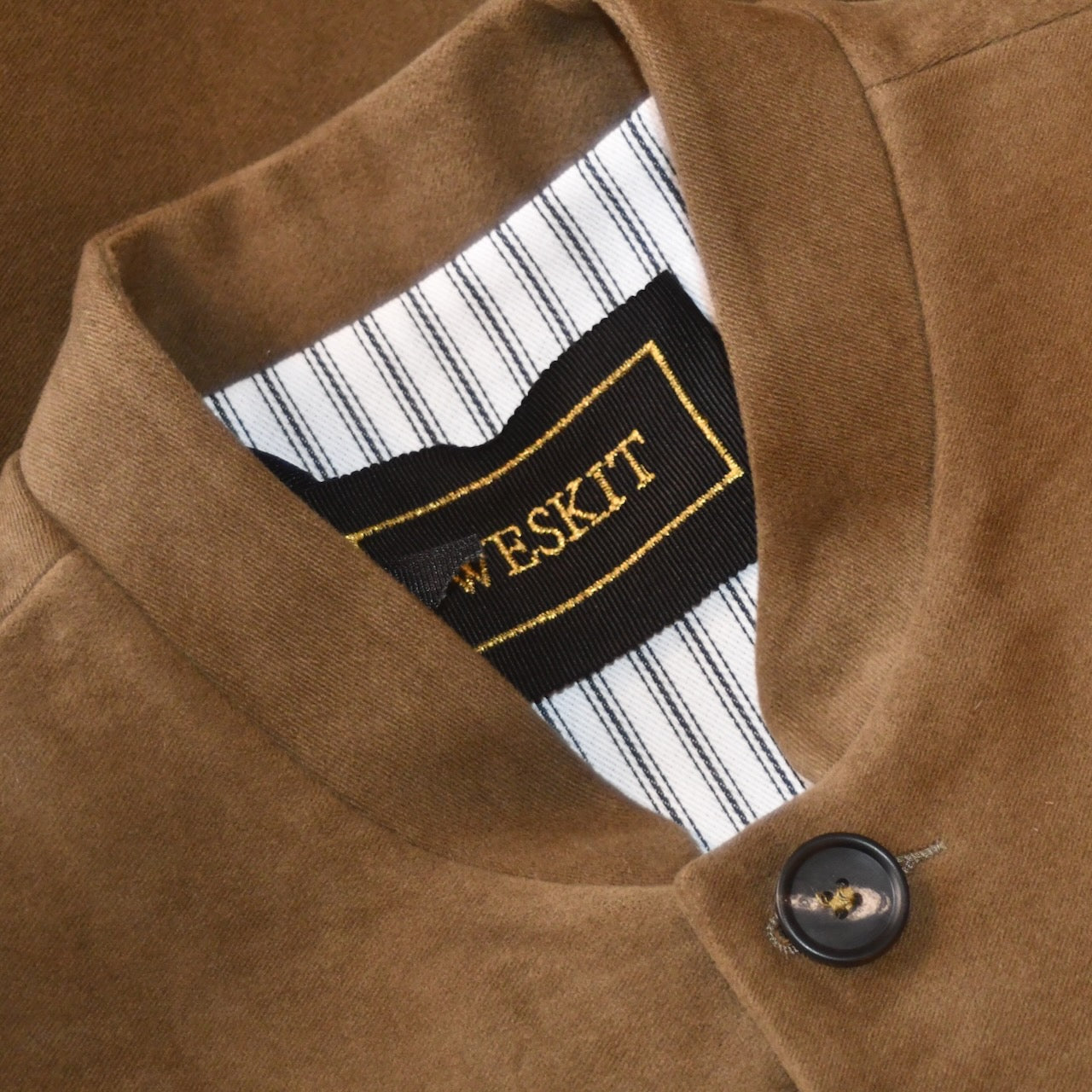 Close up of Mandarin Collar on Slim Cut Nehru Style Gilet in Khaki Moleskin. This pure cotton gilet is lined in striped cotton Ticking. With a Nehru collar and five pockets it will take you everywhere and keep you warm on cooler days. Made by Hand in London. Shown unbuttoned over yellow trousers and white shirt.