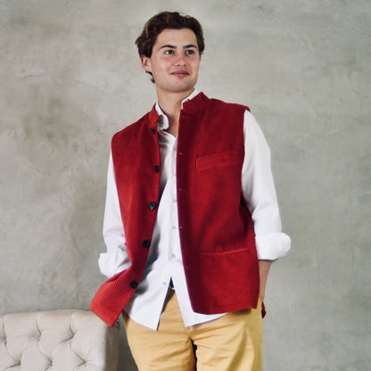 Fitted Nehru-Style Gilet in Claret Corduroy. This pure cotton corduroy gilet has five pockets, neat shaping at the waist to give a nicely tailored look and a five buttoned front. Handmade in London. Standing shot with hands in pockets.