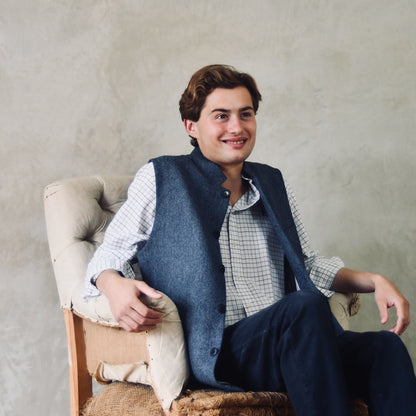 Fitted Cut Nehru-Style Gilet in Denim Blue Herringbone Tweed. A Tailored Cut waistcoat in magical mid-to-warmer weight lambswool tweed with a herringbone patterning.
Giving the overall effect of a delightful denim from a distance, this textiles brightens up on closer inspection as the depths and vivid colours of its patterning are revealed.
Smart, elegant and urbane – but as easy to match with jeans as it is to wear more formally. Seated View.