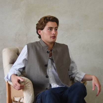 Nehru-style fitted gilet in relaxed brown Irish linen. Handmade in London this slim-cut waistcoat has a Nehru collar, is fully lined in pure cotton ticking and has five pockets. Seated unbuttoned shot.
