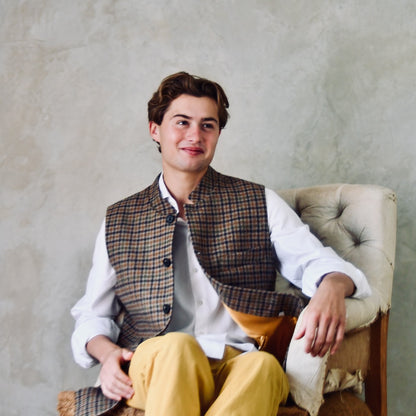 A superbly fine tweed that Nehru Style Gilet. Worth every penny, it will take you everywhere, smartly. A smokey brown and dull green weave a stunning check against a warm hued background, creating an unusual and bold check that will hold its own anywhere. Woven in Somerset, Cu. Made and Finished in London, this Gilet is entirely made in Great Britain. Seated Model Shot.