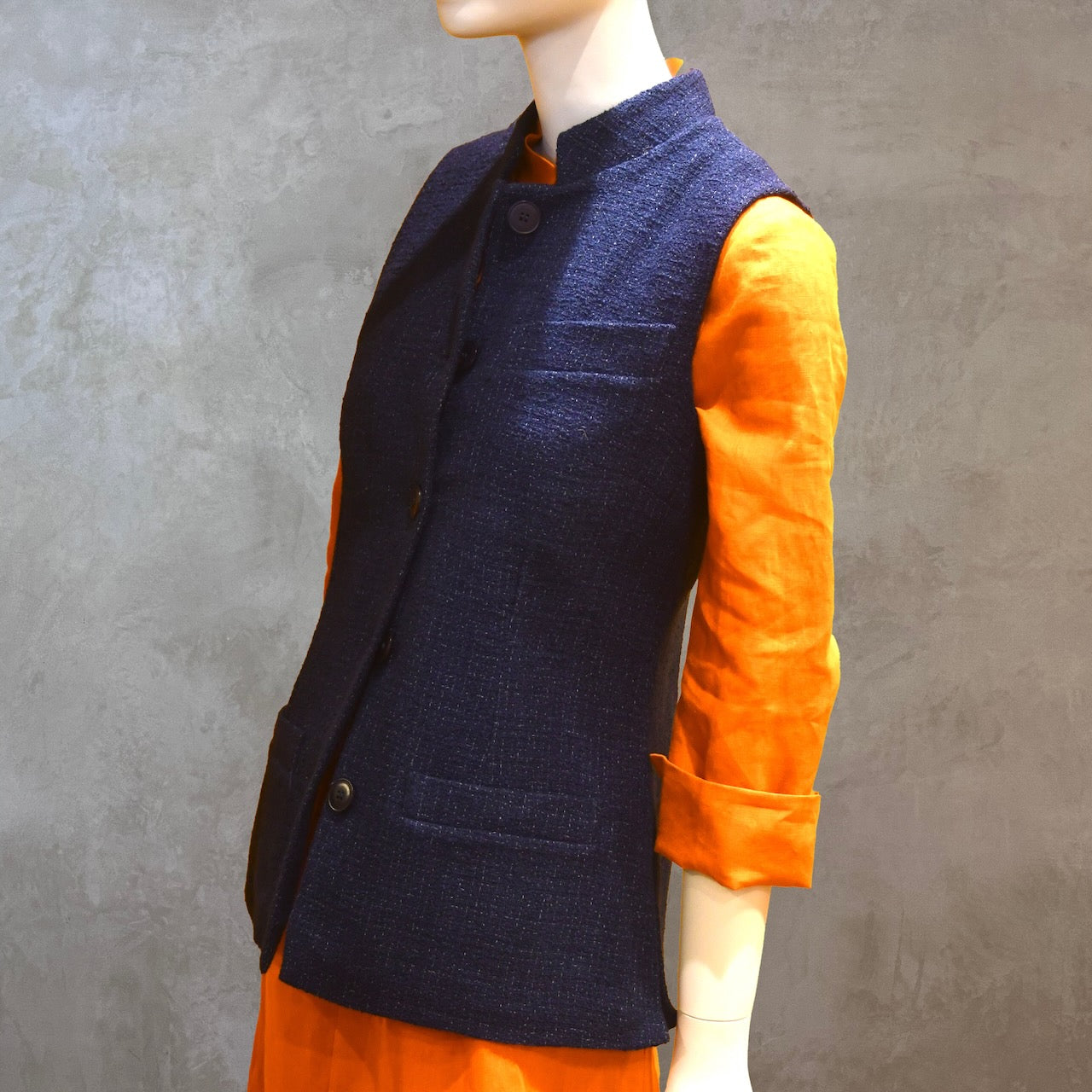Ladies Shaped Gilet in Linton Tweeds boucle. A deep navy spring-summer weight fabric, this gilet packs superbly and can be worn everywhere. Handmade in London. Side Shot shown over orange lightweight linen dress from EdNerat.