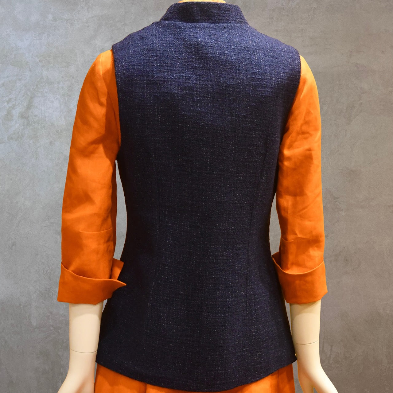 Ladies Shaped Gilet in Linton Tweeds boucle. A deep navy spring-summer weight fabric, this gilet packs superbly and can be worn everywhere. Handmade in London. Back Shot shown over orange lightweight linen dress from EdNerat.
