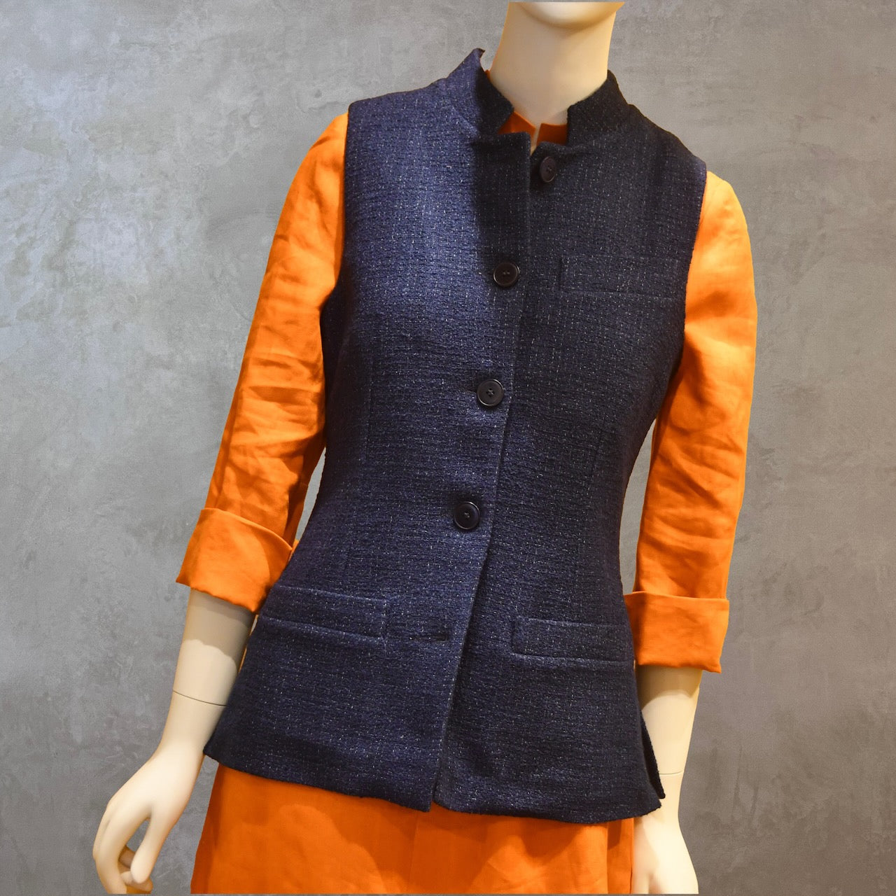Ladies Shaped Gilet in Linton Tweeds boucle. A deep navy spring-summer weight fabric, this gilet packs superbly and can be worn everywhere. Handmade in London. Full Length Buttoned Front Shot shown over orange lightweight linen dress from EdNerat.