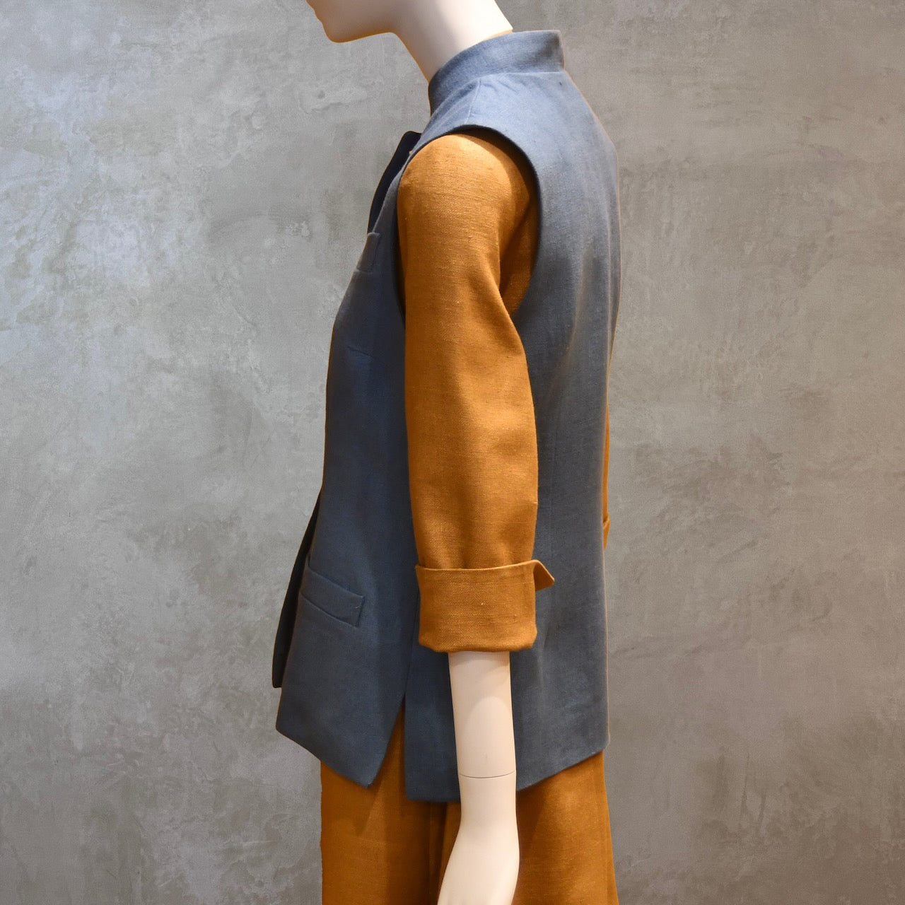 Side on Shot of Ladies Gilet in Light Blue Irish Linen. Handmade in London this waistcoat has a Nehru collar, cotton ticking lining, bust darts as well as front and rear waist-shaping darts. Shown over mustard coloured EdNerat dress.