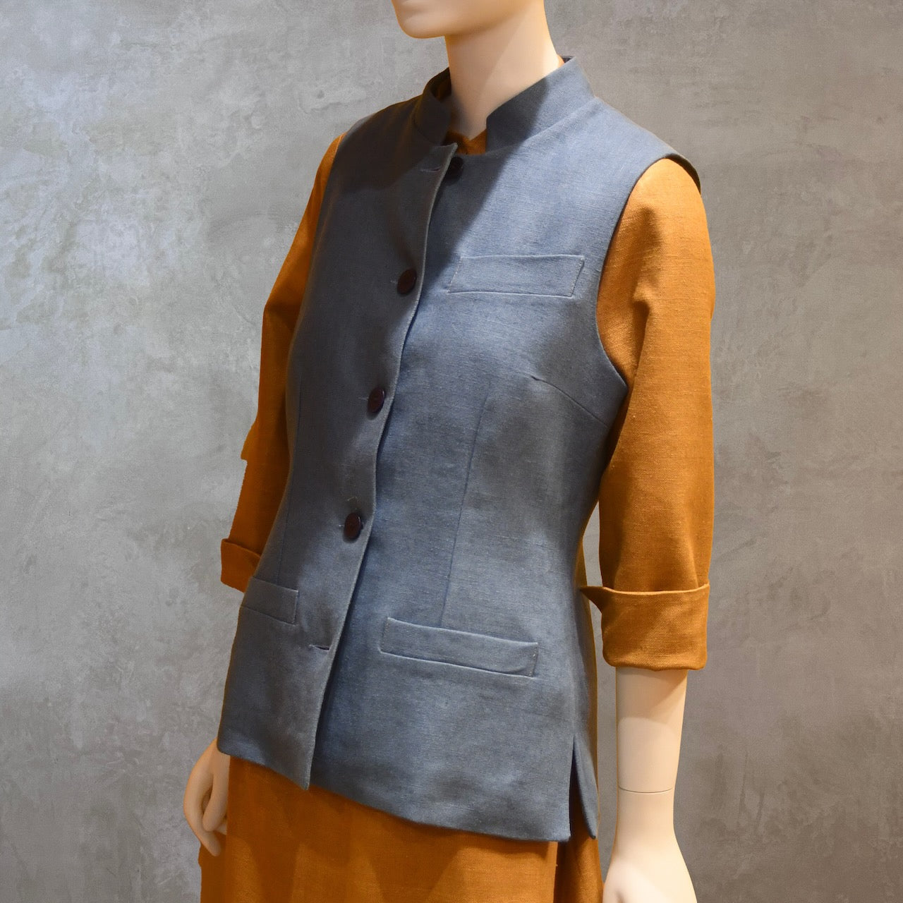 Angled Shot of Ladies Gilet in Light Blue Irish Linen. Handmade in London this waistcoat has a Nehru collar, cotton ticking lining, bust darts as well as front and rear waist-shaping darts. Shown over mustard coloured EdNerat dress.