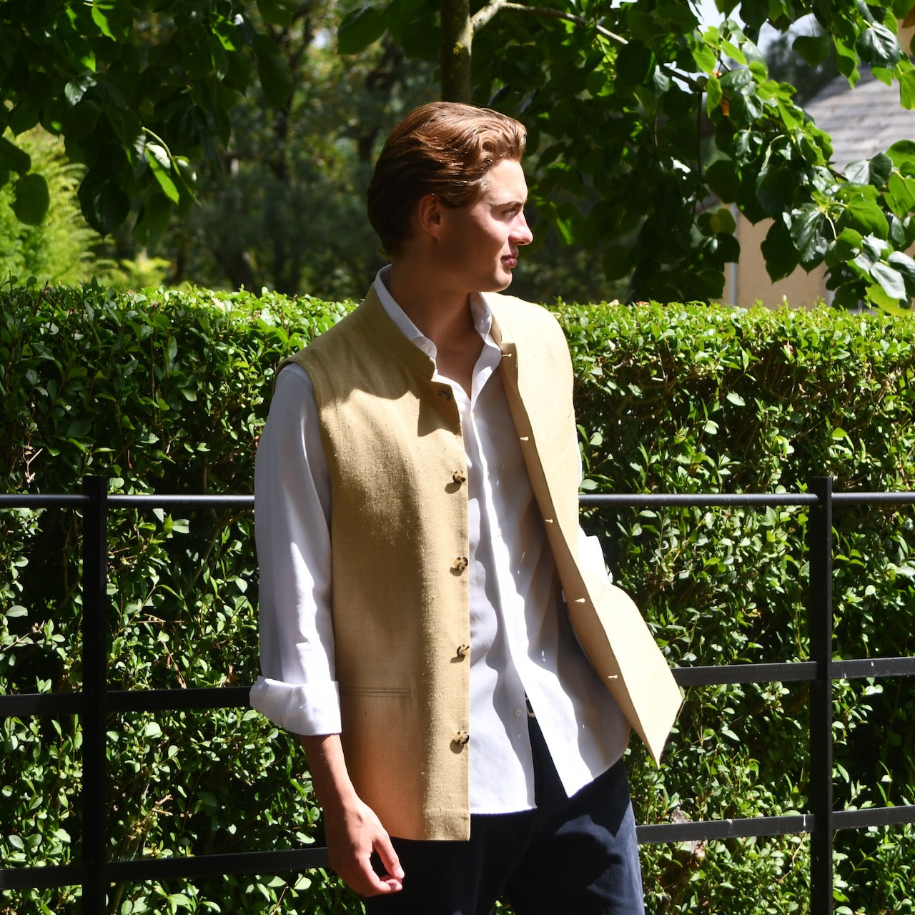 Sand Coloured Silk Matka Gilet. Handmade in London. Photographed in Wales. Shown against Estate Fencing with luscious green backdrop. Side View.