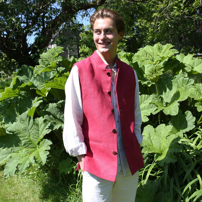 Red Linen Nehru Style Gilet. Handmade in London. Entirely made in Great Britain. Shown outside over white linen shirt and trousers against leafy green backdrop.