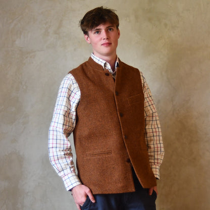 Bespoke Waistcoat in pure wool tweed in russet brown. Handmade in London, this Nehru-style gilet is shown worn buttoned up over a checked shirt and blue chino trousers.
