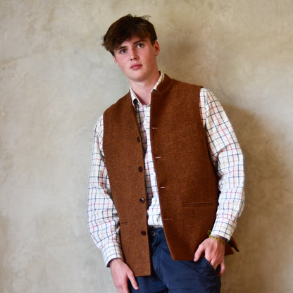 Bespoke Waistcoat in pure wool tweed in russet brown. Handmade in London, this Nehru-style gilet is shown worn open over a checked shirt and blue chino trousers.