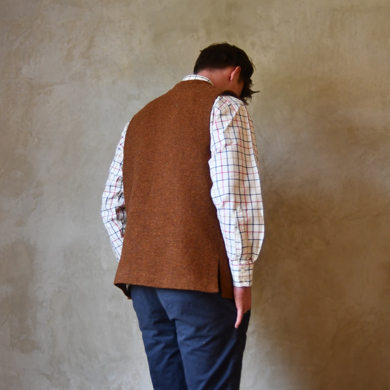 Rear view of Bespoke Waistcoat in pure wool tweed in russet brown. Handmade in London, this Nehru-style gilet is shown worn over a checked shirt and blue chino trousers.