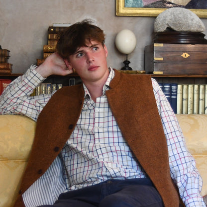 Bespoke Waistcoat in pure wool tweed in russet brown. Handmade in London, this Nehru-style gilet is shown seated and worn open over a checked shirt and blue chino trousers.