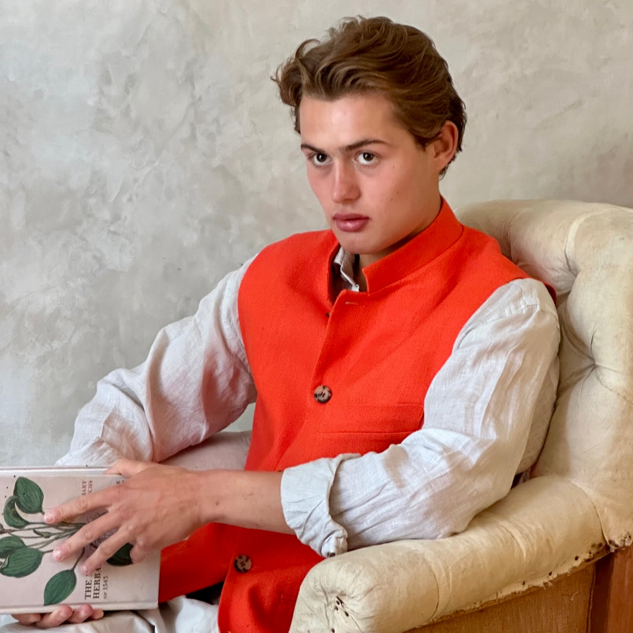 Slim fit nehru style gilet in summer weight silk weave fabric. Handmade in London, this orange silk matka fitted waistcoat has a Nehru collar, Courtney & Co buttons and is fully lined in striped cotton ticking. Seated Shot.