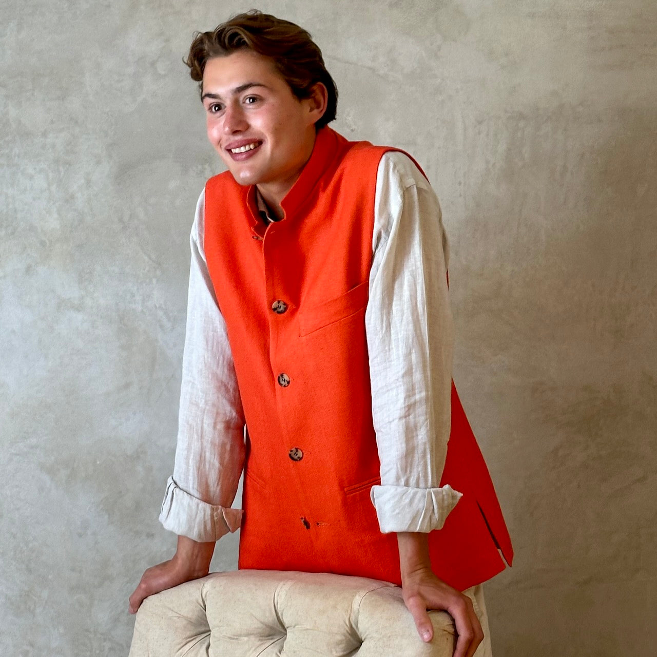 Slim fit nehru style gilet in summer weight silk weave fabric. Handmade in London, this orange silk matka fitted waistcoat has a Nehru collar, Courtney & Co buttons and is fully lined in striped cotton ticking. Standing Buttoned Shot with Chair.