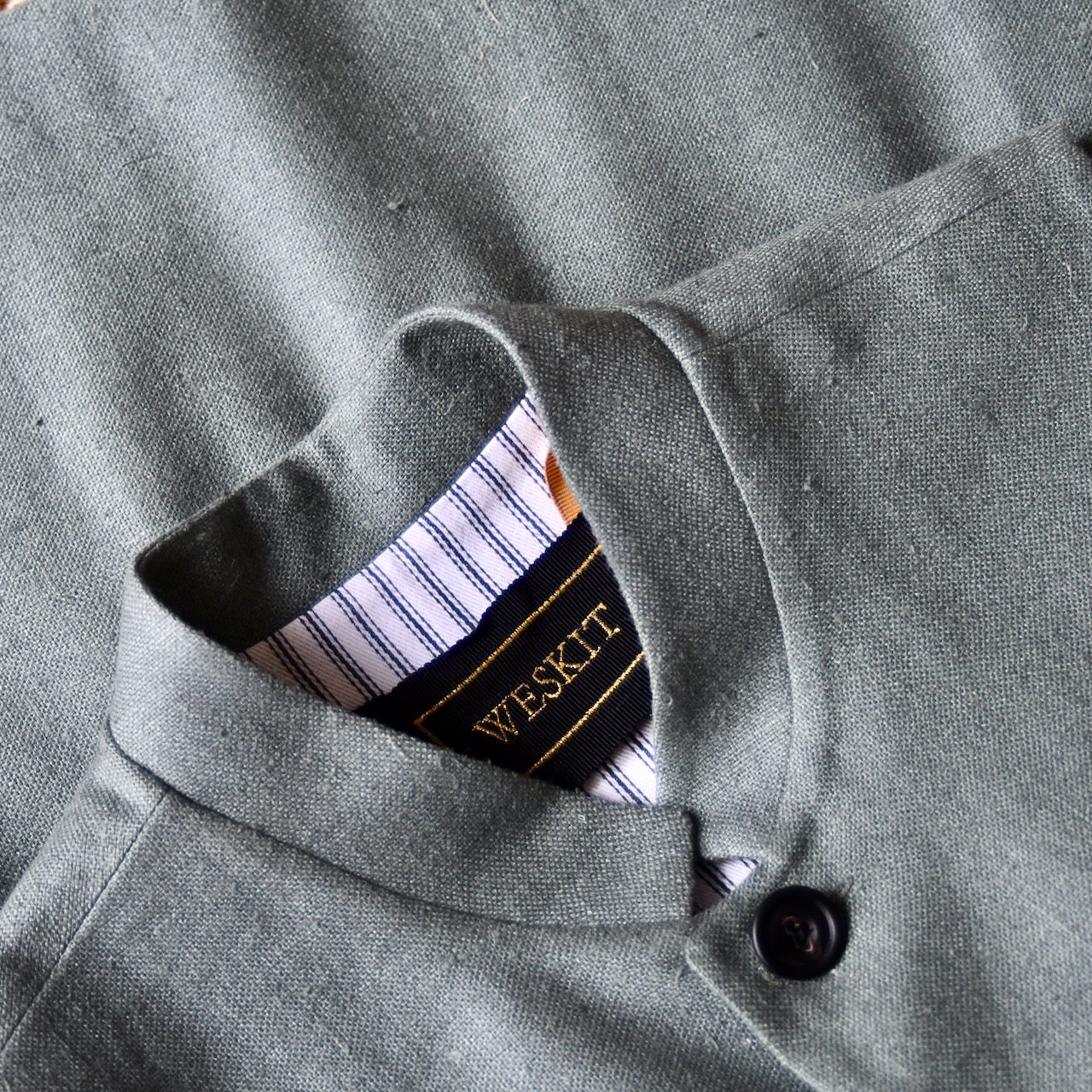 Close up on collar of slim cut summer weight nehru style gilet in pale green silk weave. Handmade in London this fitted waistcoat has a Nehru  collar and is fully lined in cotton ticking. Real horn buttons. Shot outside in summer.