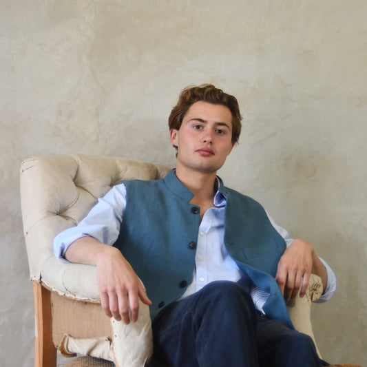 Light blue Slilm cut linen gilet from Weskit. Handmade in London, this slubby linen waistcoat is made in pure Irish linen and lined in striped cotton ticking. Mandarin collar and Courtney and Co buttons. Entirely made in Great Britain. Seated View.