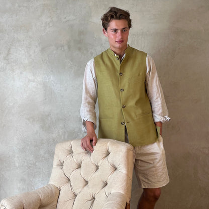 Relaxed fit summer weight gilet in green silk weave. Handmade in London this gilet has a Mandarin collar and is fully lined in pure cotton ticking. Made in Great Britain. Model standing next to chair.