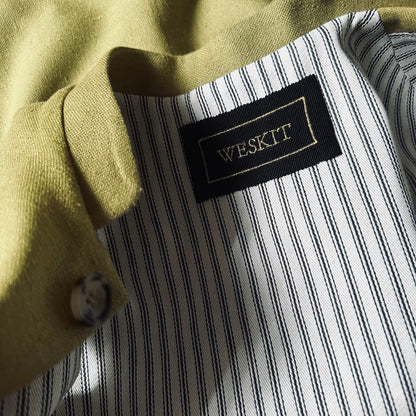 Relaxed fit summer weight gilet in green silk weave. Handmade in London this gilet has a Mandarin collar and is fully lined in pure cotton ticking. Made in Great Britain. Close up shot showing mandarin collar, black silk embroidered label and striped cotton ticking lining.