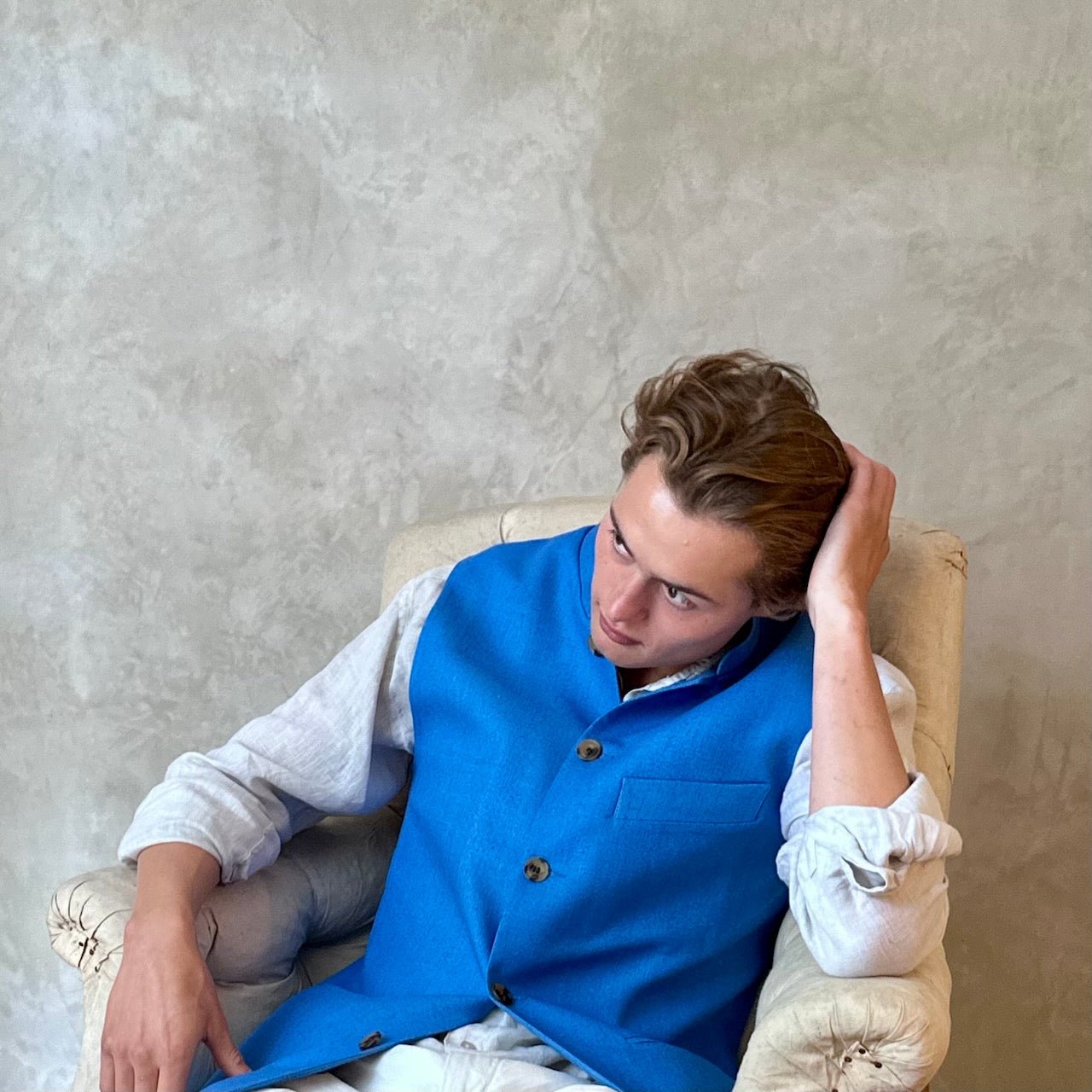 Slim fit gilet in sky blue summer weight silk weave. Handmade in London this fitted waistcoat has five pockets and a Nehru collar. Made in Great Britain. Seated side shot.