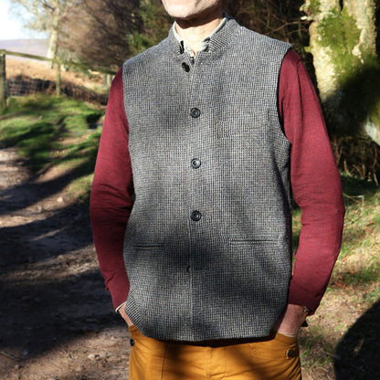 Warm Nehru Style Gilet in pure wool tweed from Hawick. Handmade in London this waistcoat has a gentle patterning in blue and shades of taupe and grey. Ample pocket space, side vents and a nead Mandarin Collar. Outside shot showing gilet worn over red jumper.