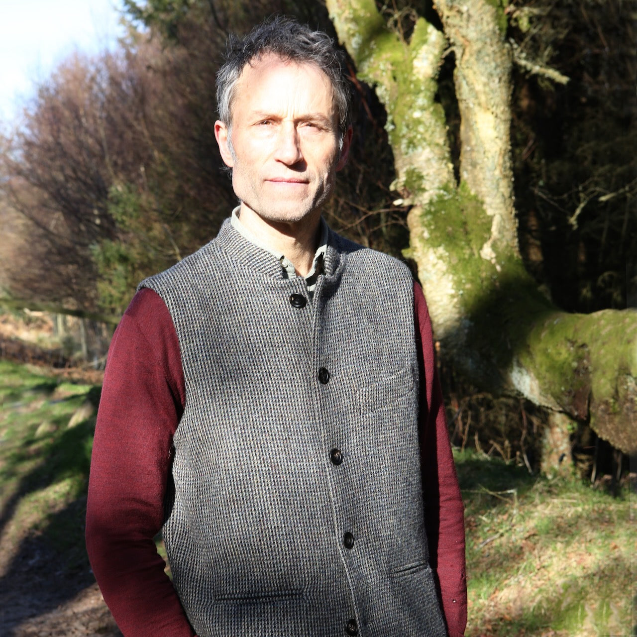 Warm Nehru Style Gilet in pure wool tweed from Hawick. Handmade in London this waistcoat has a gentle patterning in blue and shades of taupe and grey. Ample pocket space, side vents and a nead Mandarin Collar. Outside shot against woodland.