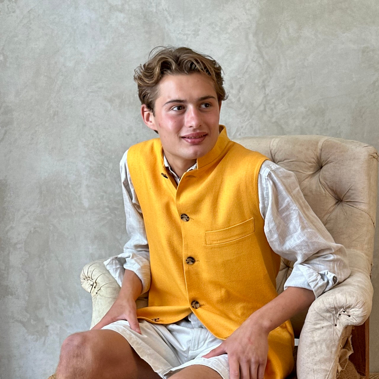 Seated model wearing summer weight slim-fit gilet in bright yellow silk weave. Handmade in London this tailored cut waistcoat has a Mandarin Collar, Striped Cotton Ticking Lining and five buttons to the front. Made in Great Britain.